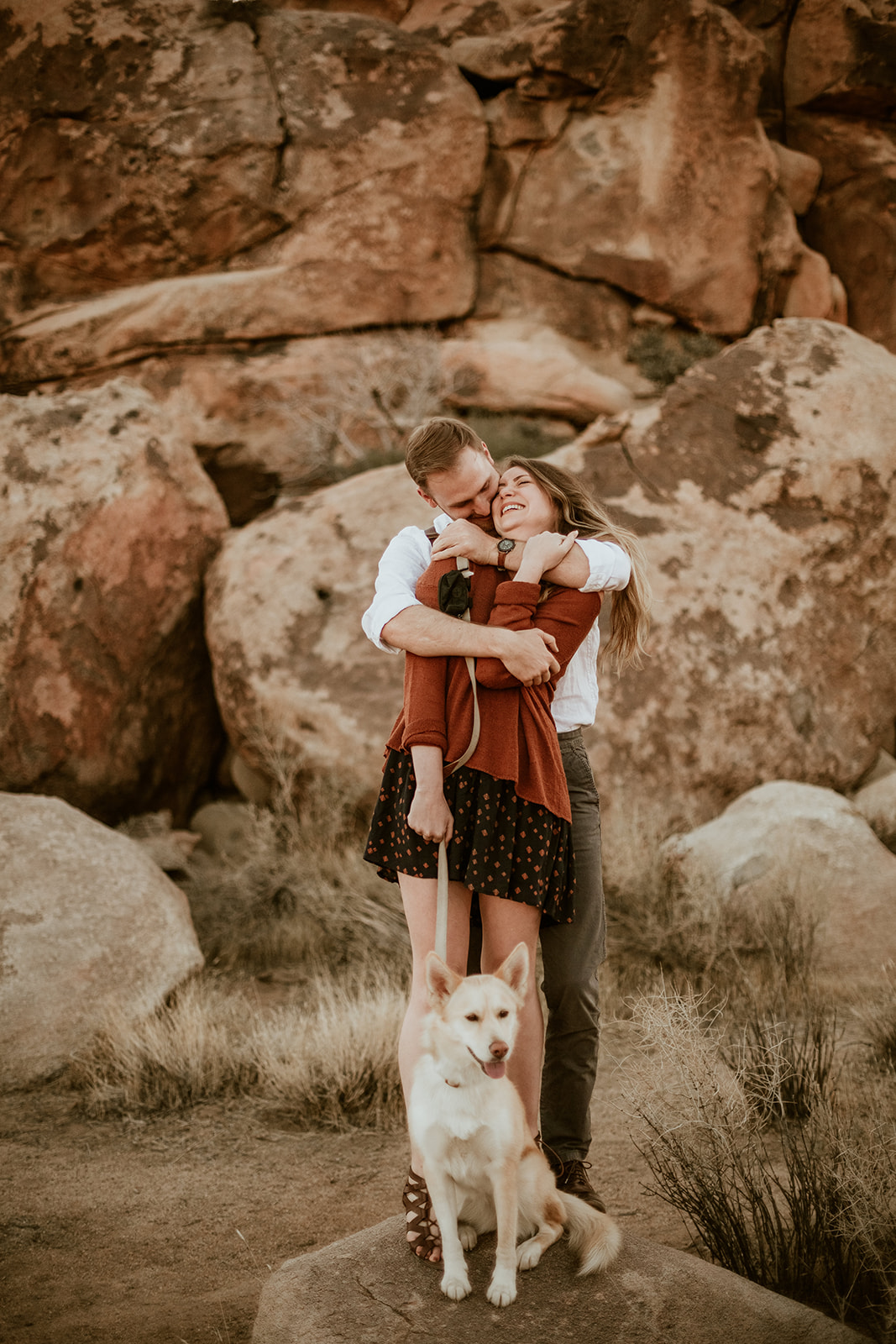 A couple holding each other with their dog among the desert boulders
