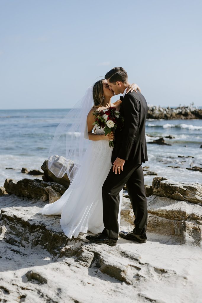 A bride and groom embrace and touch foreheads on rocky sand beside the ocean, with the bride holding a bouquet of flowers, under a clear blue sky.