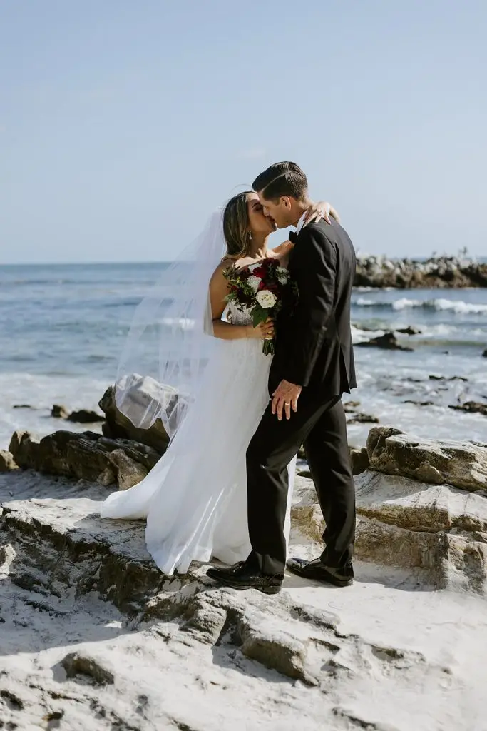 A bride and groom embrace and touch foreheads on rocky sand beside the ocean, with the bride holding a bouquet of flowers, under a clear blue sky.