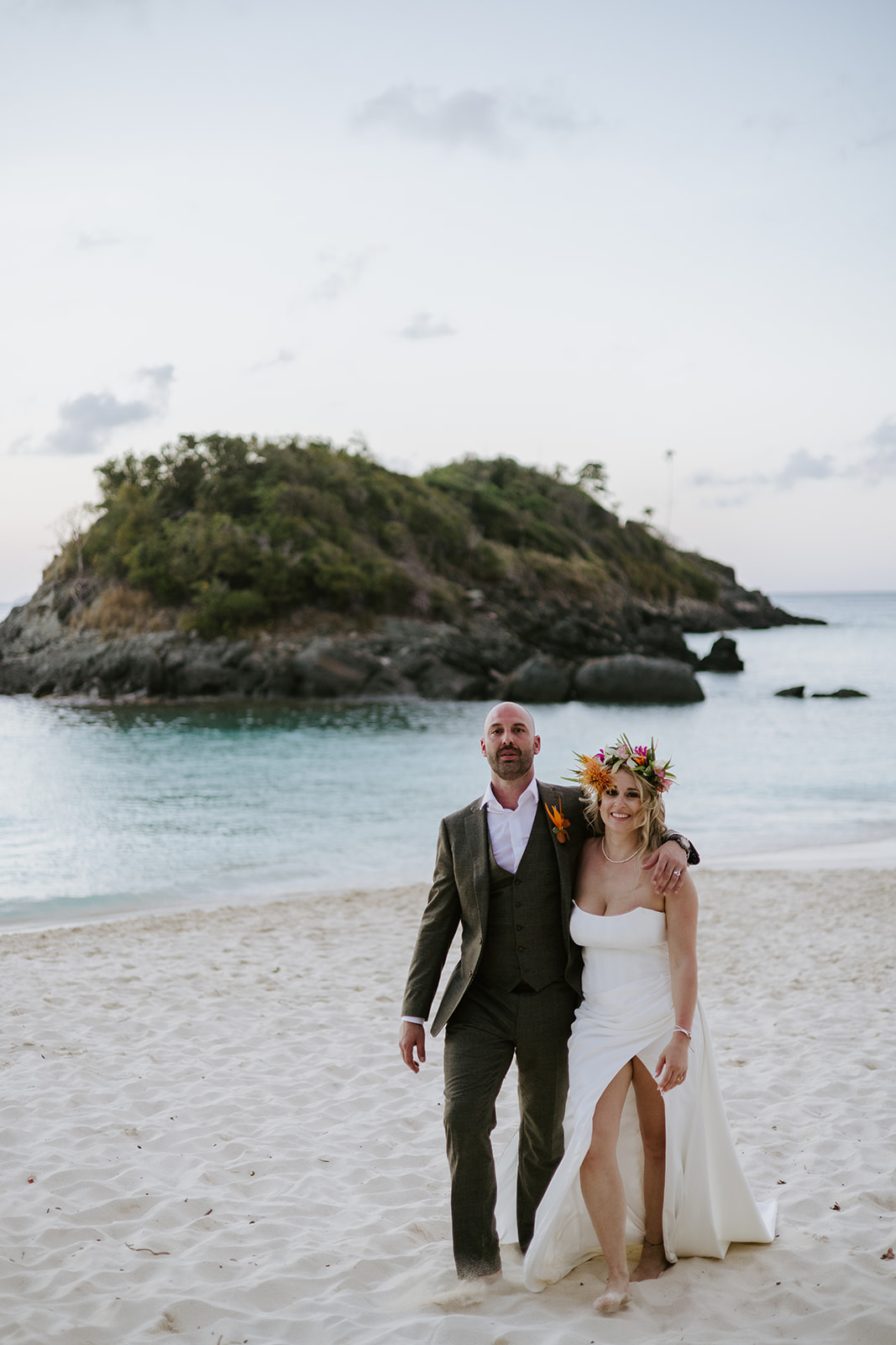 A couple stands on a sandy beach at sunset, dressed in wedding attire. The woman wears a white dress with a floral crown, and the man wears a suit. Behind them is calm blue water and a rocky, green island.