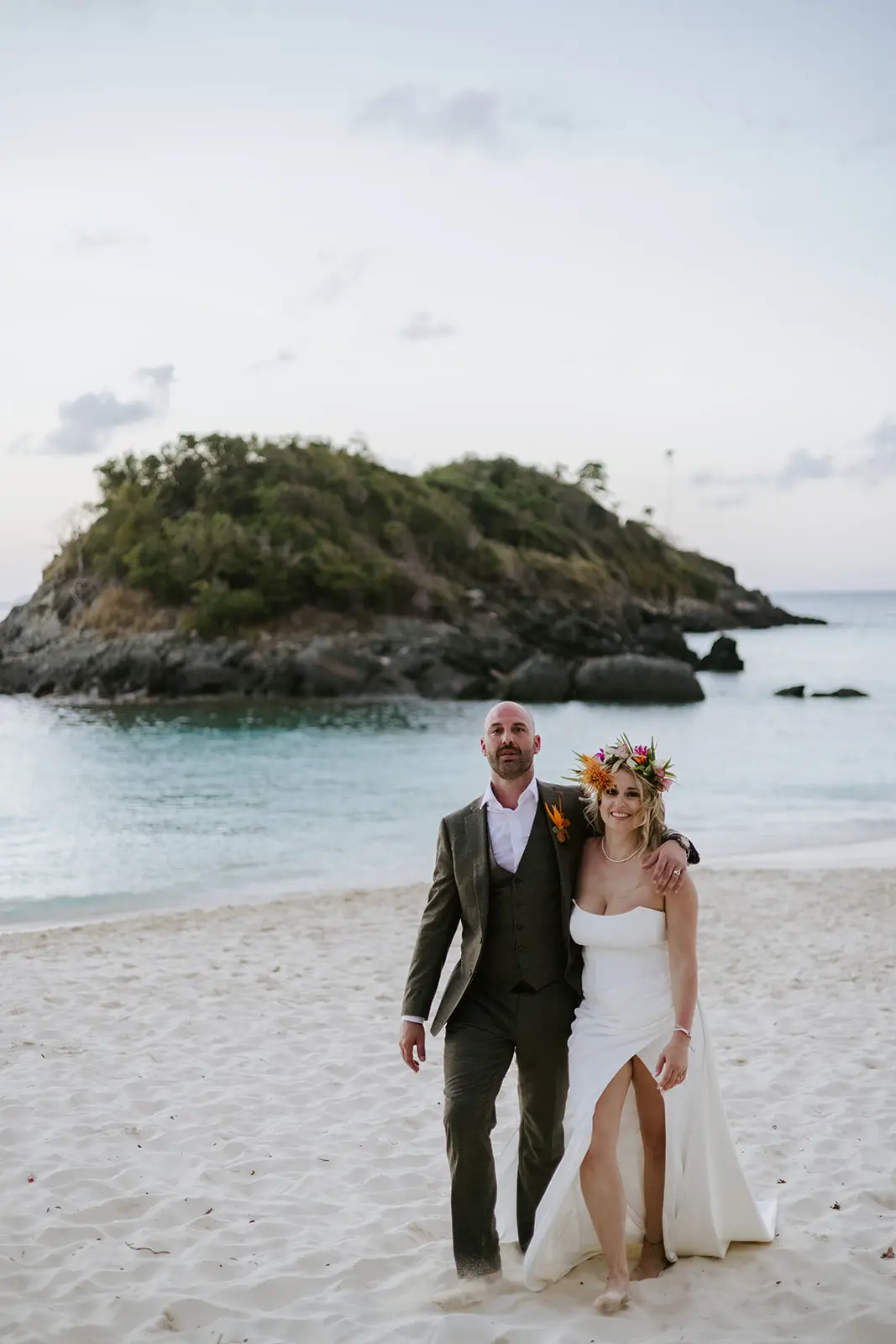 A couple stands on a sandy beach at sunset, dressed in wedding attire. The woman wears a white dress with a floral crown, and the man wears a suit. Behind them is calm blue water and a rocky, green island.