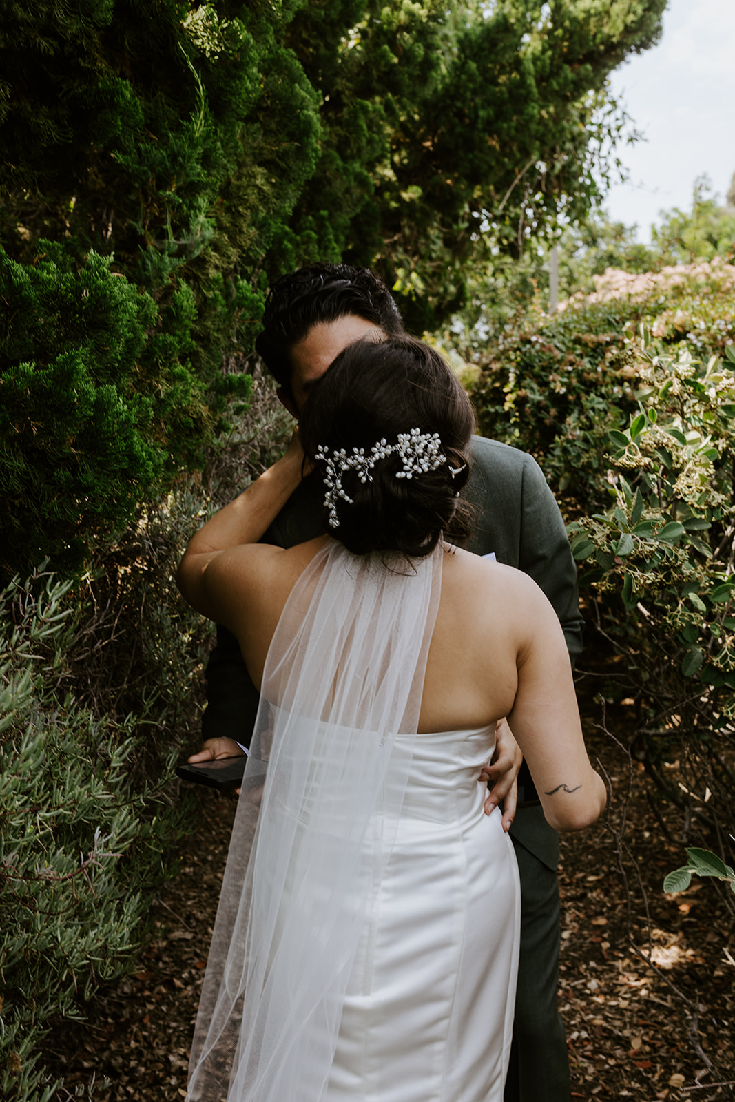 A bride in a white dress and veil, with a pearl hairpiece, embraces and kisses a groom in a suit outdoors, surrounded by greenery and trees. The couple stands close together on a garden path.
