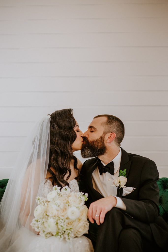 A bride and groom share a kiss while seated on a green couch. The bride wears a white gown, veil, and holds a bouquet of white flowers; the groom wears a black tuxedo with a bow tie and boutonnière.