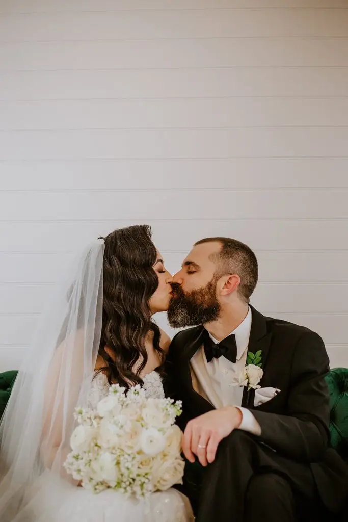 A bride and groom share a kiss while seated on a green couch. The bride wears a white gown, veil, and holds a bouquet of white flowers; the groom wears a black tuxedo with a bow tie and boutonnière.