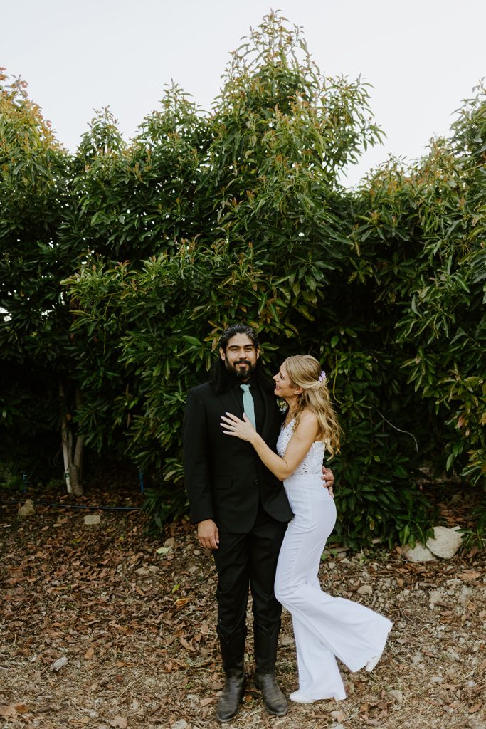 A couple poses outdoors in front of green leafy trees. The man wears a black suit with a light green tie, and the woman, in a sleeveless white dress, embraces him while smiling. The ground is covered in dry leaves.