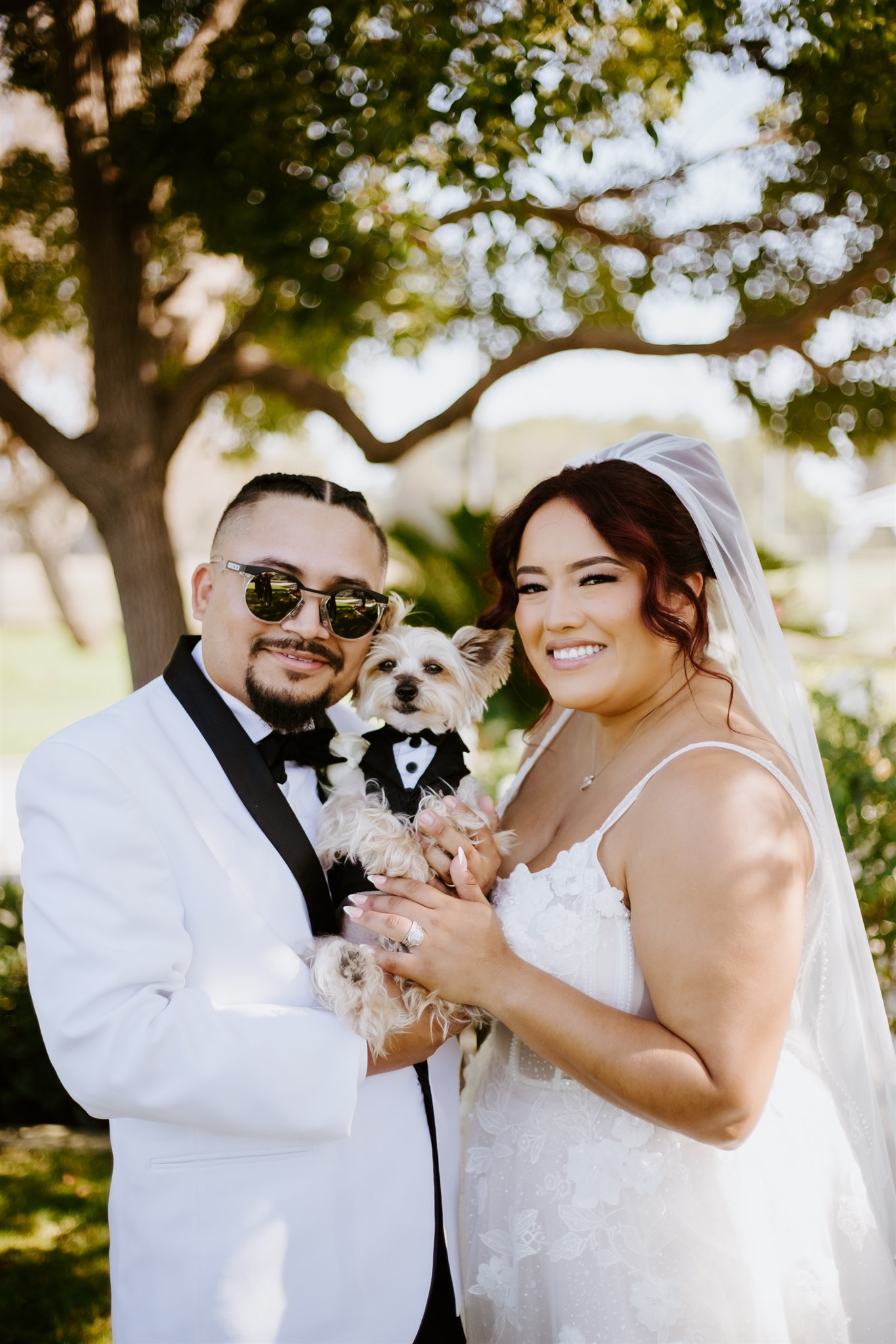 A bride and groom in wedding attire smile outdoors while holding a small dog dressed in a tuxedo. Lush green trees and sunlight are visible in the background.