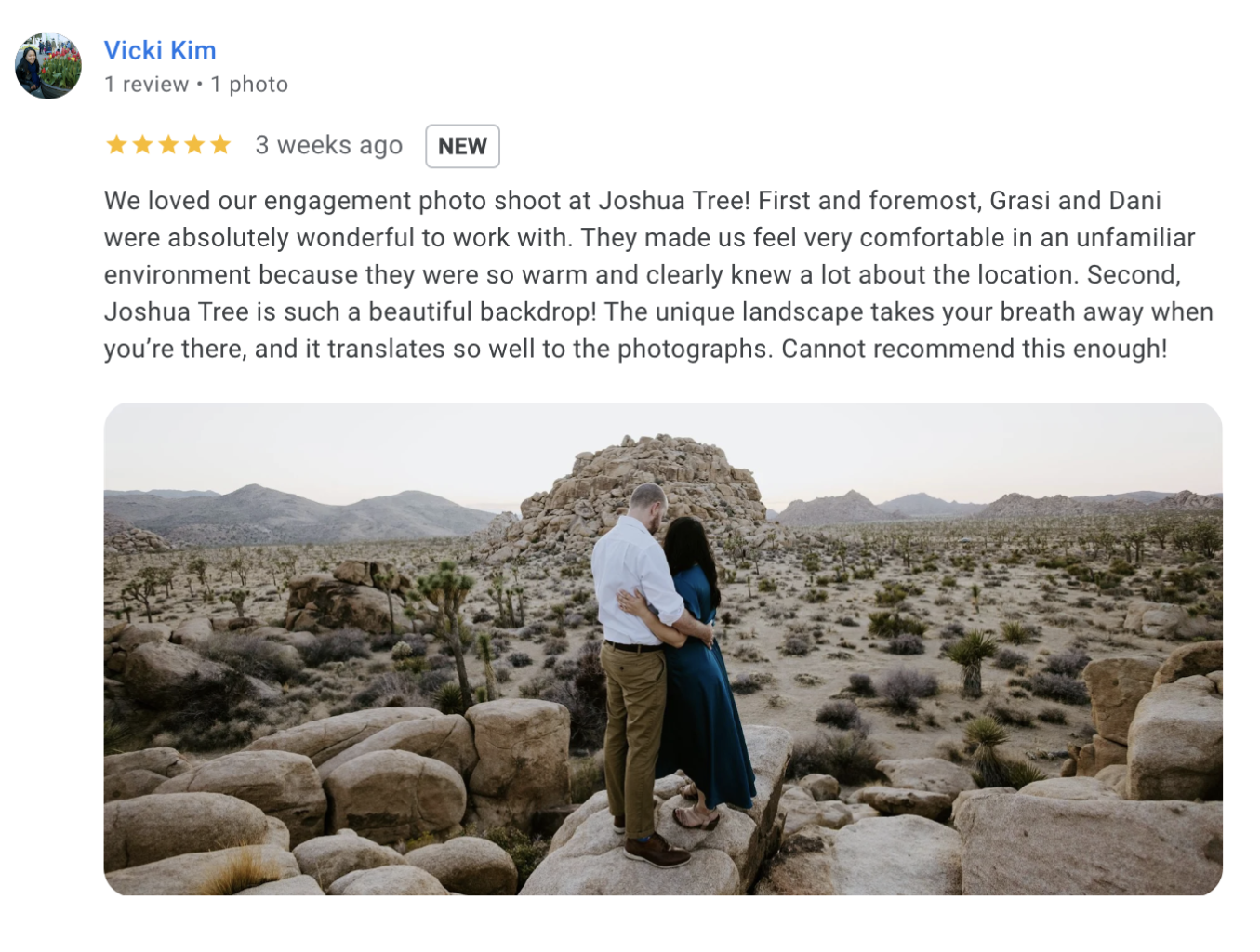 A couple embraces on a rocky area in Joshua Tree National Park, surrounded by desert plants and distant mountains under a clear sky.