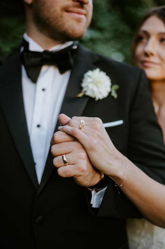 A couple in formal wedding attire holds hands close-up, showing their wedding rings. The groom wears a black tuxedo with a white boutonniere, while the brides engagement ring and delicate bracelet are visible.